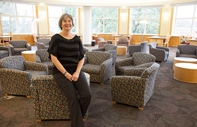 Joan Oakes, Ecampus student services specialist, sits against the back of a short cushioned chair in the OSU Valley Library. She smiles and wears a black and gray striped shirt.