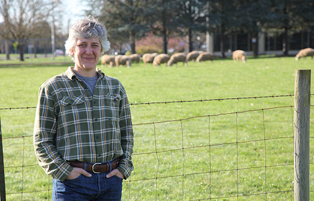 Yvette Gibson stands in front of a wire fence with sheep in the background. Yvette has her hands in the pockets of her denim jeans and wears a green plaid button-up shirt.