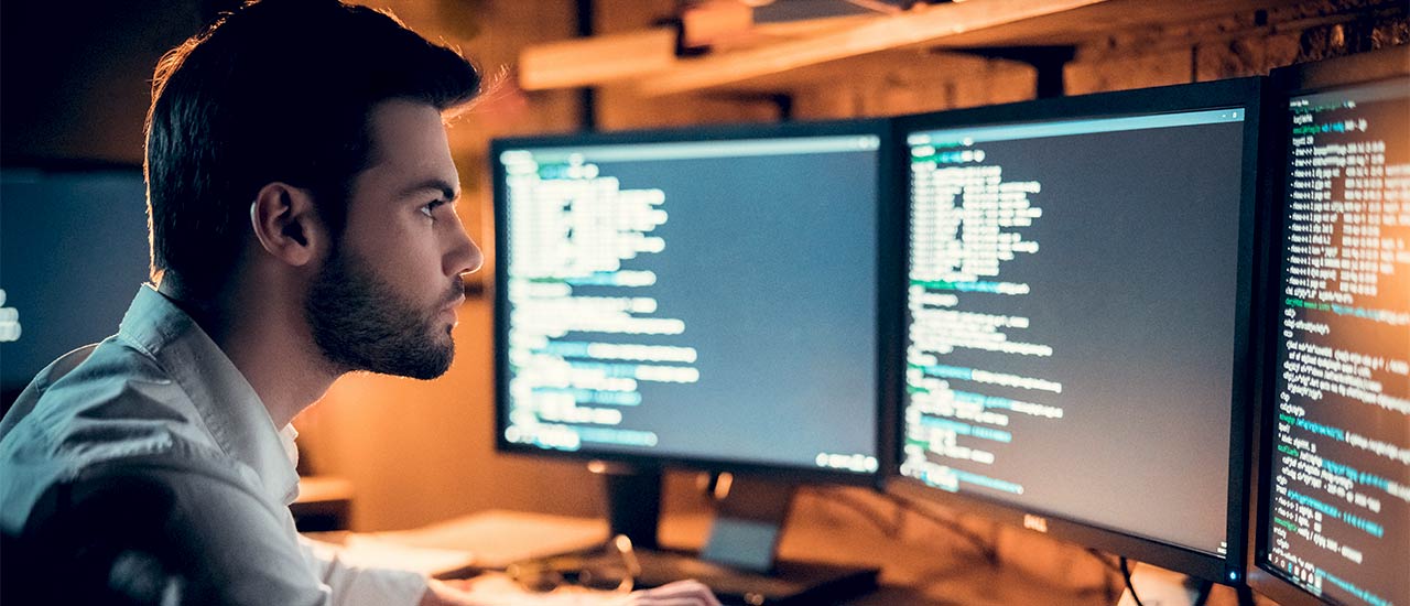 A bearded man sits in front of three computer monitors analyzing website back end code