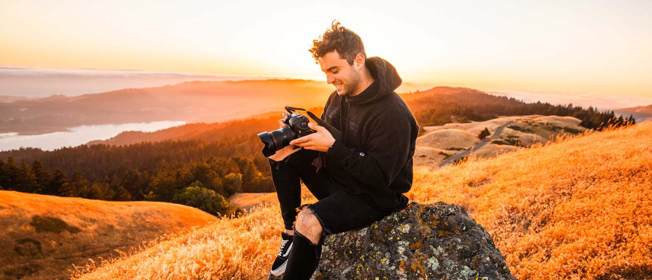 Young man sitting on hillside reviewing shot on DSLR camera with beautiful sunset behind him