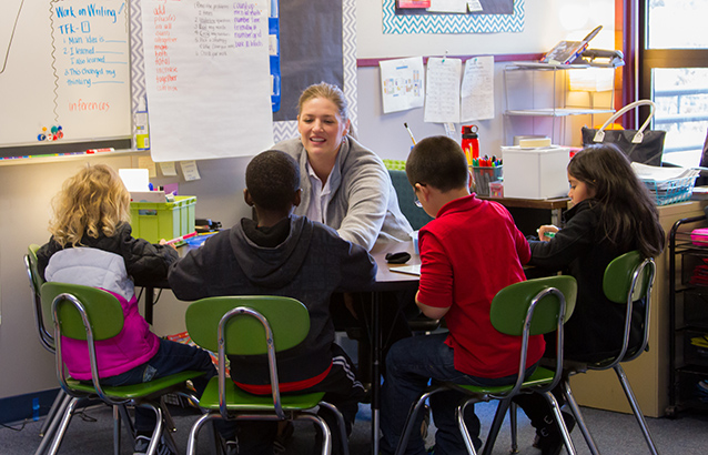 A teacher works with four students in the Beaverton School District. They all sit around a table in small green chairs.