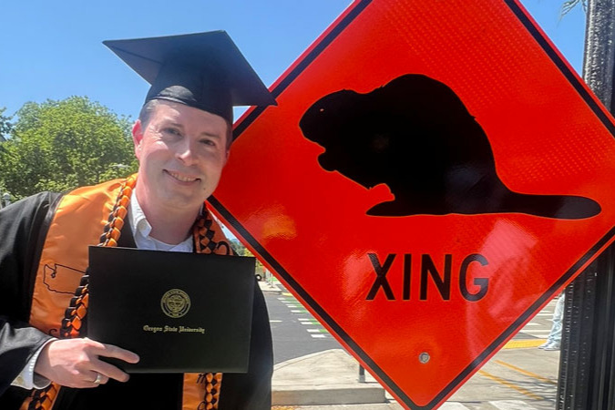 Oregon State graduate Adam Lemcio stands in a cap and gown holding a diploma next to an orange traffic sign with a beaver image.
