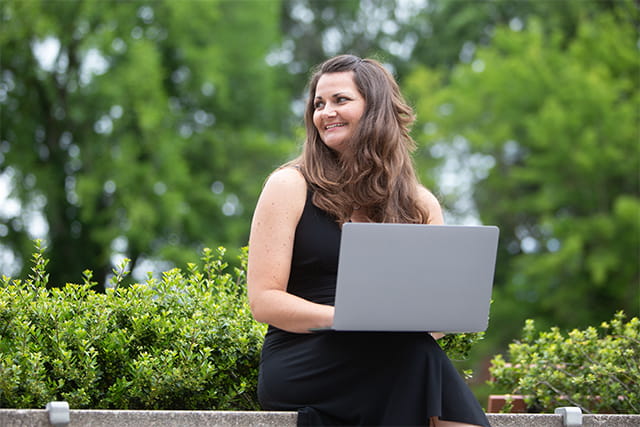 A woman sitting outdoors with a laptop on her lap, smiling and looking to the side, surrounded by greenery and trees.