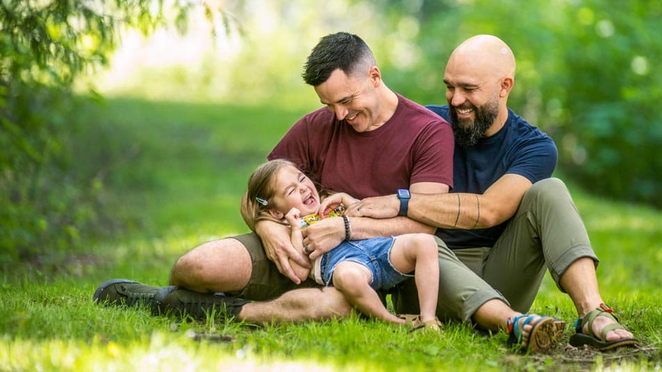 Bobby Disler, who earned a degree in Women, Gender, and Sexuality Studies through Oregon State University Ecampus, sits in a green field smiling with his husband as they hold their laughing daughter.