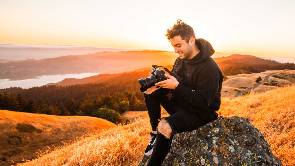 Photographer Jake Chamseddine sits on a rock atop a hill, holding a camera. Jake is a graduate of Oregon State University's digital communication arts bachelor's degree program.