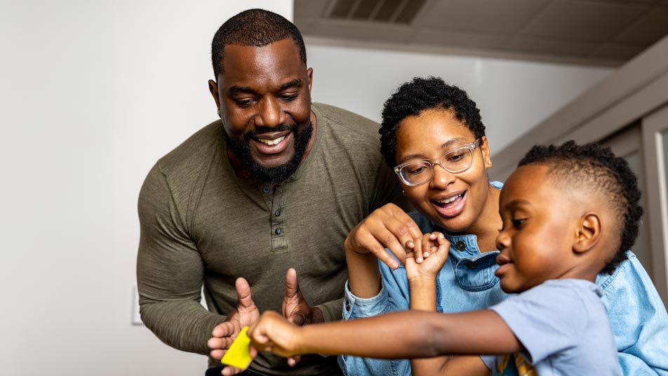 Ravonne Byrd, an Oregon State University computer science graduate, sits at a table playing with her son while her husband stands clapping behind them.