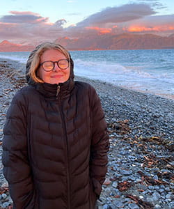 Tann Wise wearing a dark puffer jacket standing on a rocky beach with ocean waves and a mountain range at sunset.
