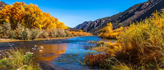 Clear river winding through rocky banks and golden autumn trees beneath mountain peaks and a bright blue sky