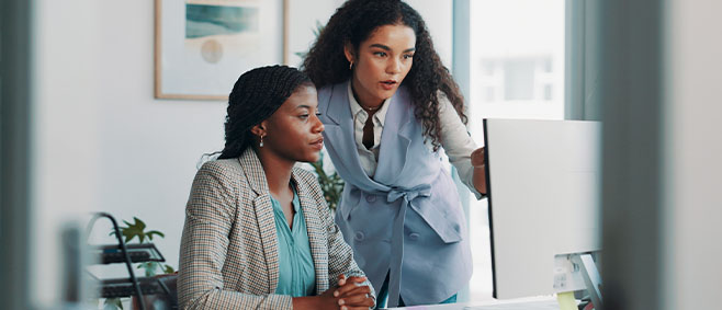 Colleague leans over a desk to review information on a computer screen with a seated IT professional.