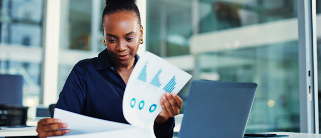 Accountant reviewing printed financial reports and charts while working at a desk with a laptop.