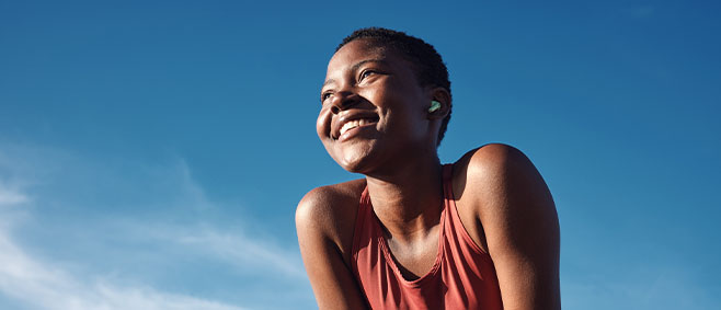 A woman in a sleeveless athletic top leans forward with arms braced on her knees, photographed from a low angle against a clear blue sky.