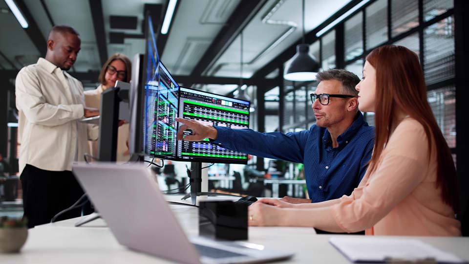 People working together at a desk while reviewing data displayed on multiple monitors.