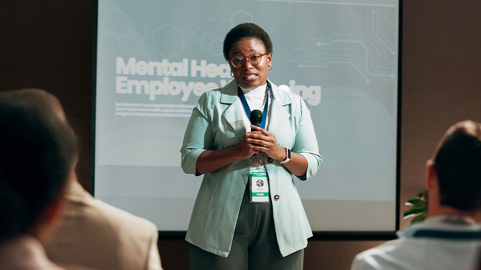 A person wearing a lanyard holds a microphone in front of a room of listeners. The person is standing in front of a screen that says, “Mental health employee screening.”