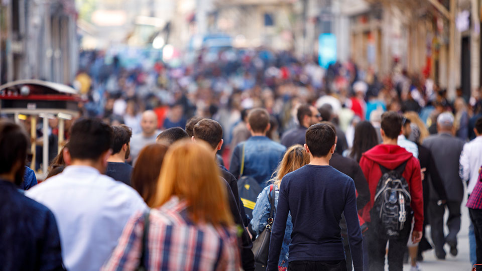 A very busy sidewalk is bustling with a diverse crowd of people dressed for a moderate day as far as the camera can see