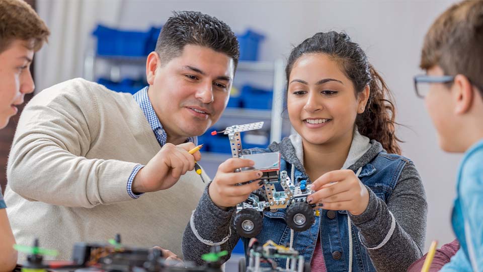 A male teacher works with a group of students on building a rudimentary hand-held robot. A young girl holds the device.