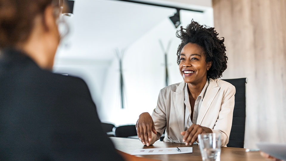 A person sits at a table across from another person in a brightly lit room. The person is smiling while talking.