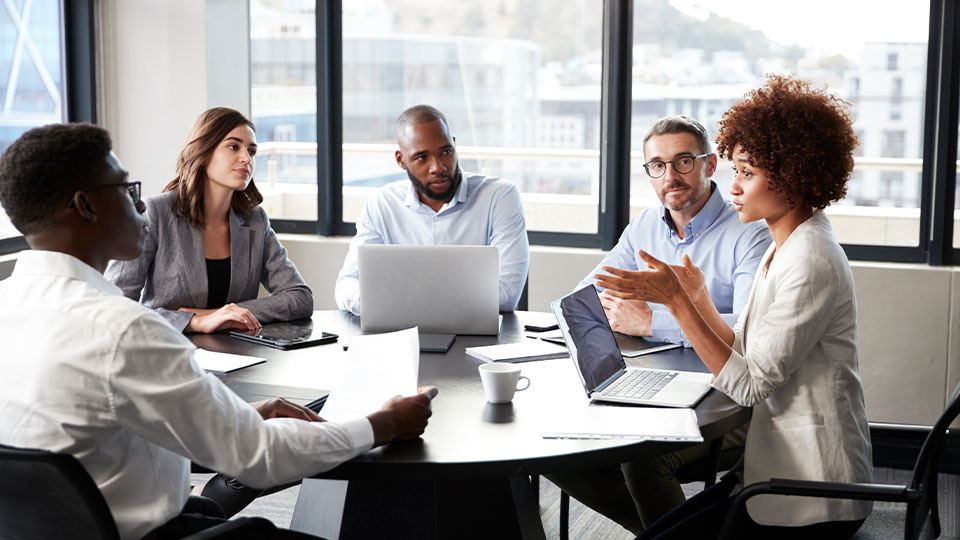 A diverse group of five people sits around a meeting table with laptops and tablets listening to a female colleague