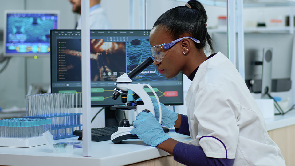 A person in a laboratory looks through a microscope while wearing protective googles and gloves. Test tubes are nearby, and computers showing data and molecules are on the desk.