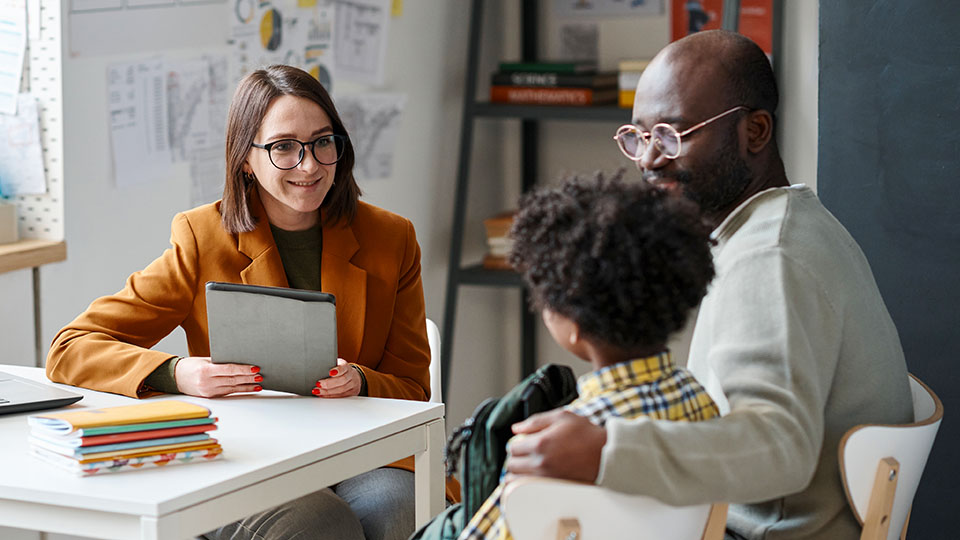 A school counselor in an office meets with a parent and a student in an office. The counselor holds paperwork while smiling at the student.