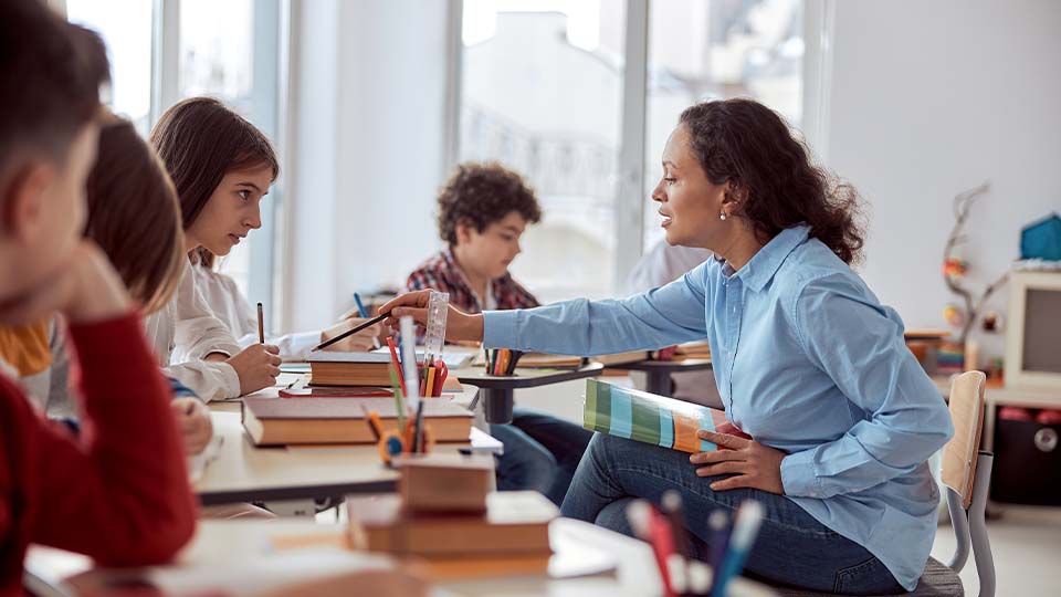 A teacher in a blue shirt leans in to guide a young student, gesturing toward her paper while others focus on their work.