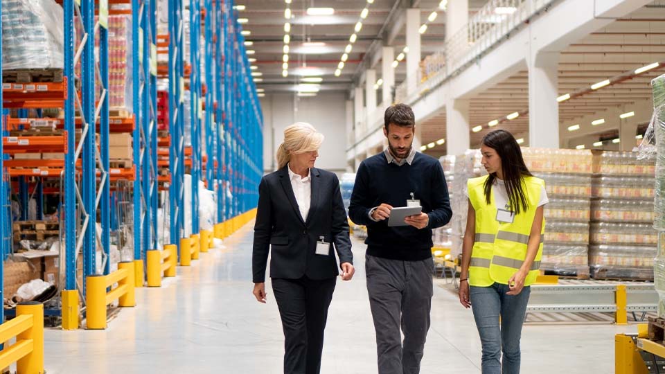 Group moving through a large warehouse with tall shelving, discussing information on a handheld device.