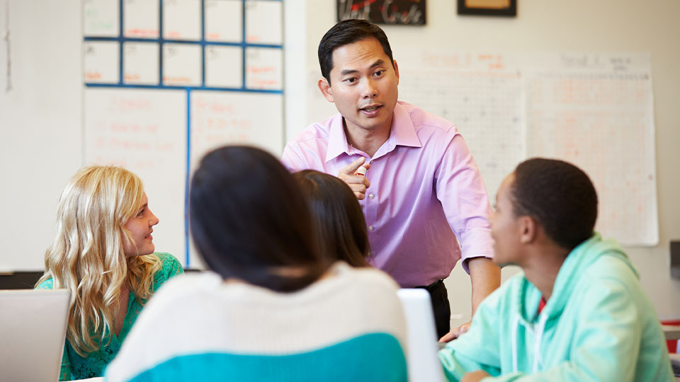 A teacher stands in front of a classroom of adult students. The teacher is talking and pointing while the students smile and listen.