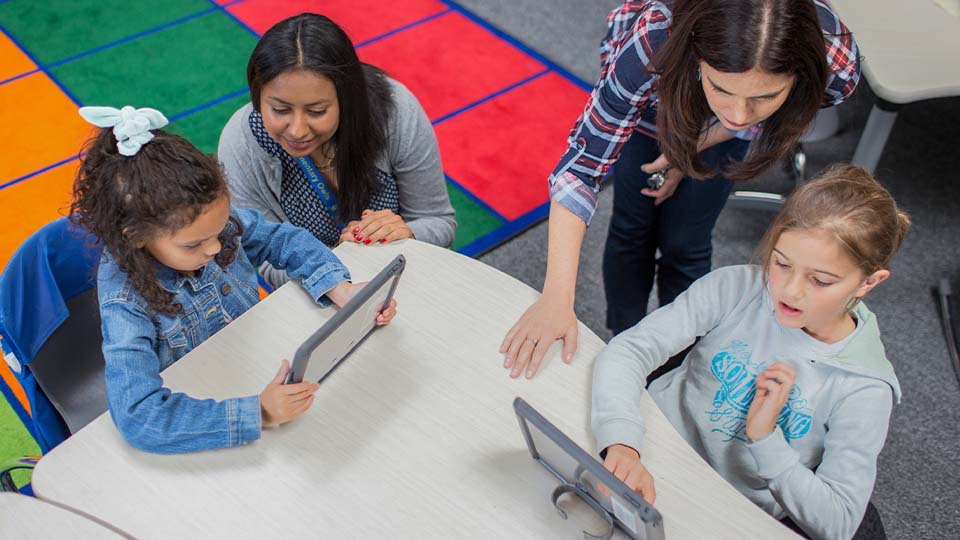 Teacher assisting young students seated around a classroom table as they use tablet devices, with a colorful rug in the background.