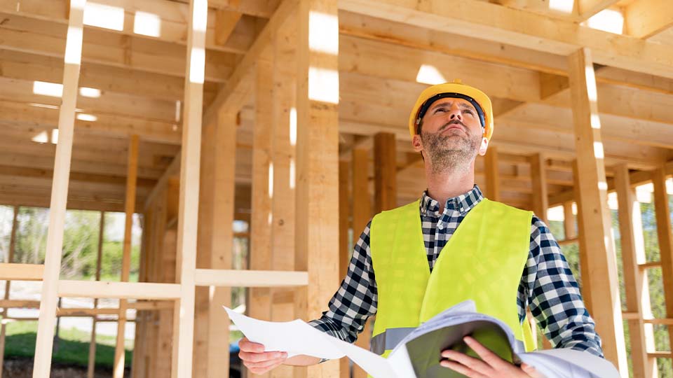 Construction professional in a hard hat and safety vest examines a wooden structure, comparing details with architectural documents.