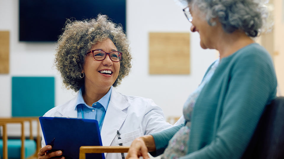 Healthcare professional holding a tablet while discussing health information with an older patient in a clinical setting.