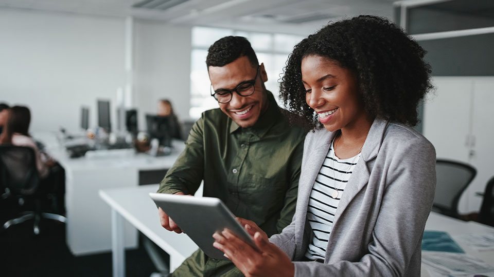 Two professionals smiling as they review information together on a digital tablet in an office.