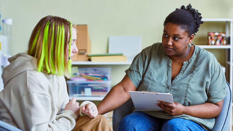 A counselor holding a clipboard attentively listens to a young person with bright green highlights in their hair, offering support in a casual, light-filled office space.