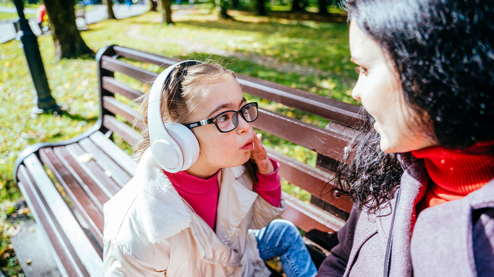 A child with Down syndrome wearing headphones and glasses gestures while sitting on a sunny park bench next to an attentive woman.