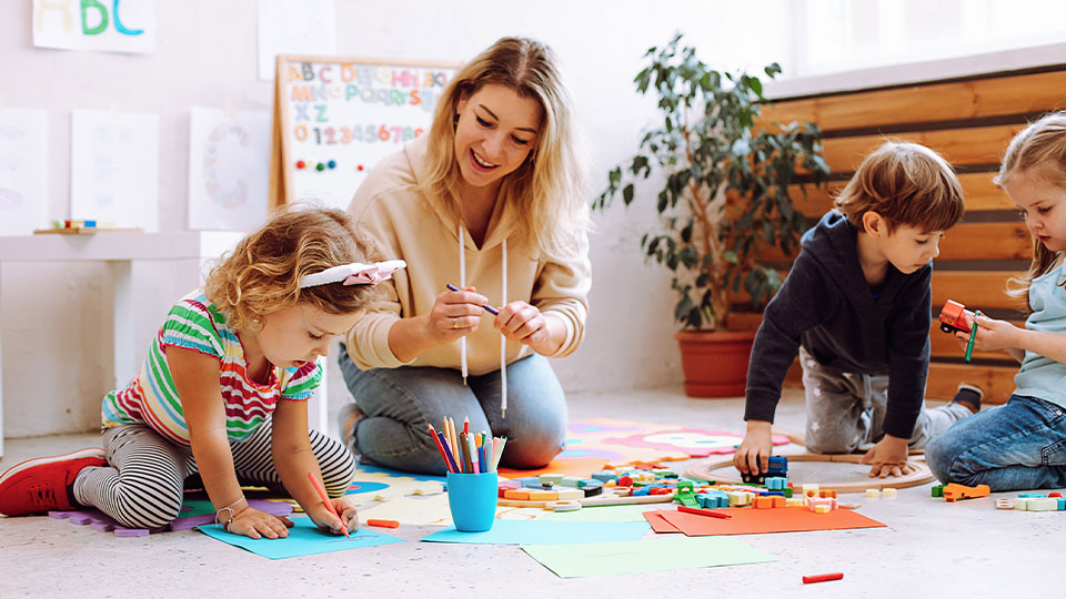 Adult sitting on the floor with three young children engaged in arts and crafts, surrounded by colorful paper, markers, and building blocks in a bright classroom setting.