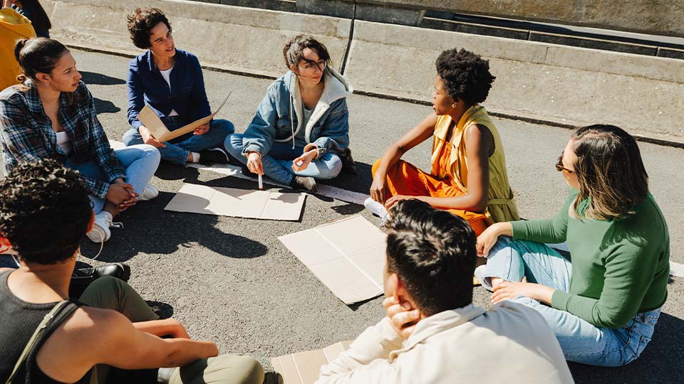 Group of people sitting in a circle outdoors, discussing and sketching ideas for a community-focused social impact project.