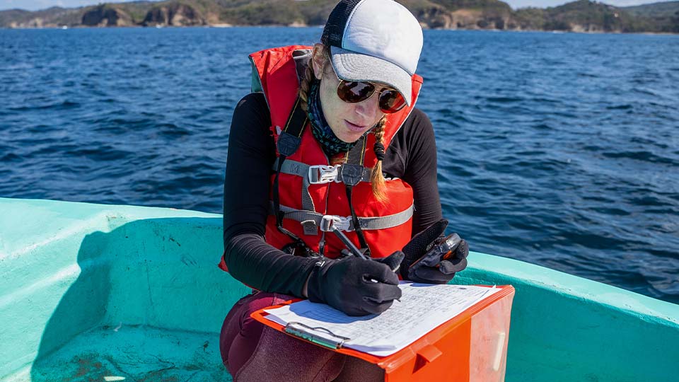 Scientist in a life jacket writes field notes on a clipboard while seated in a boat on open water.