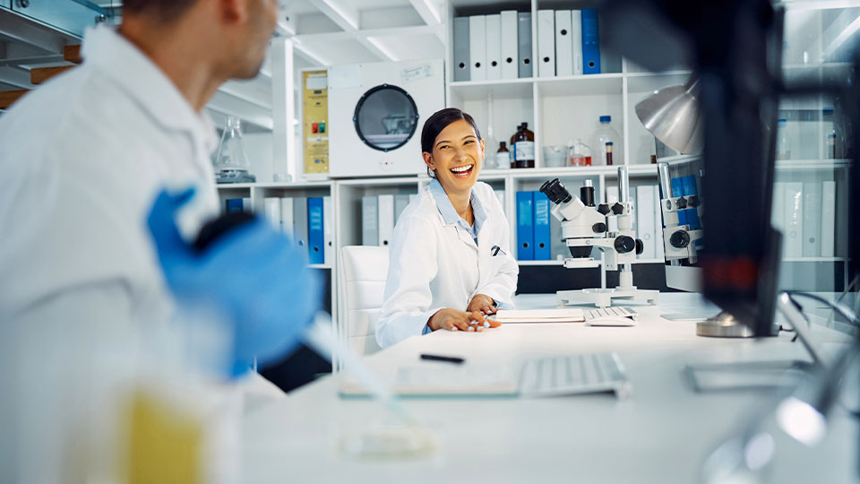 Two people in lab coats working in a modern laboratory, with one holding a pipette and the other seated near microscopes and computer equipment on a white workbench.