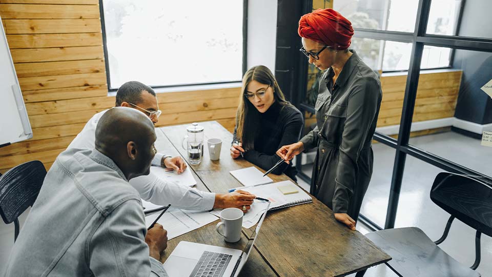 Four people gathered around a wooden table in a modern office, reviewing documents and notes, with one person standing and pointing at a notebook while others sit and write.