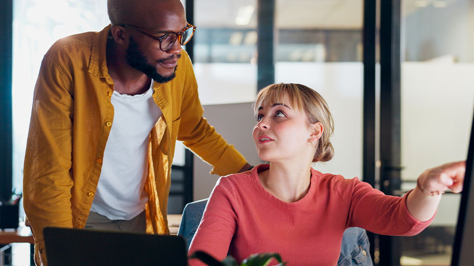 Two people collaborating in a modern office; one is standing beside a desk while the other is seated and pointing at a computer monitor.
