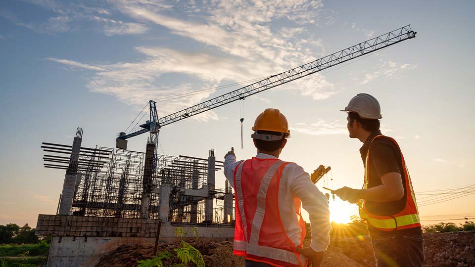 Two civil engineers in safety vests and hard hats review a construction site with cranes and a steel framework at sunset.