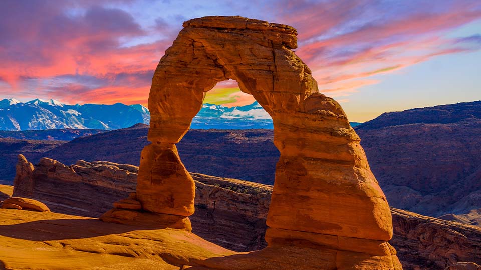 A natural sandstone arch formation in a desert landscape, known as Delicate Arch, stands prominently under a vivid sunset sky with shades of pink, orange, and purple.
