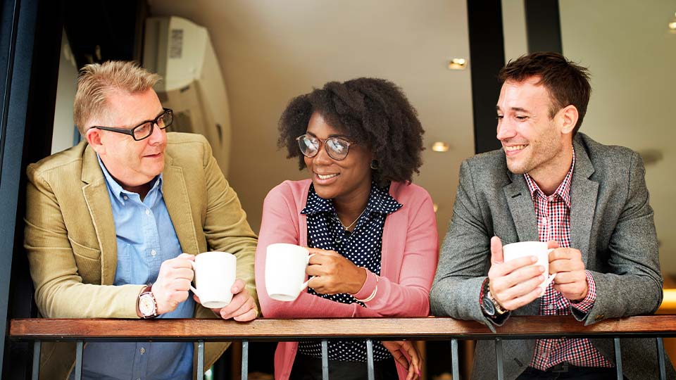 Three people standing at a railing indoors, each holding a white coffee mug. They are dressed in business casual clothing, including blazers and sweaters, and appear to be engaged in conversation.