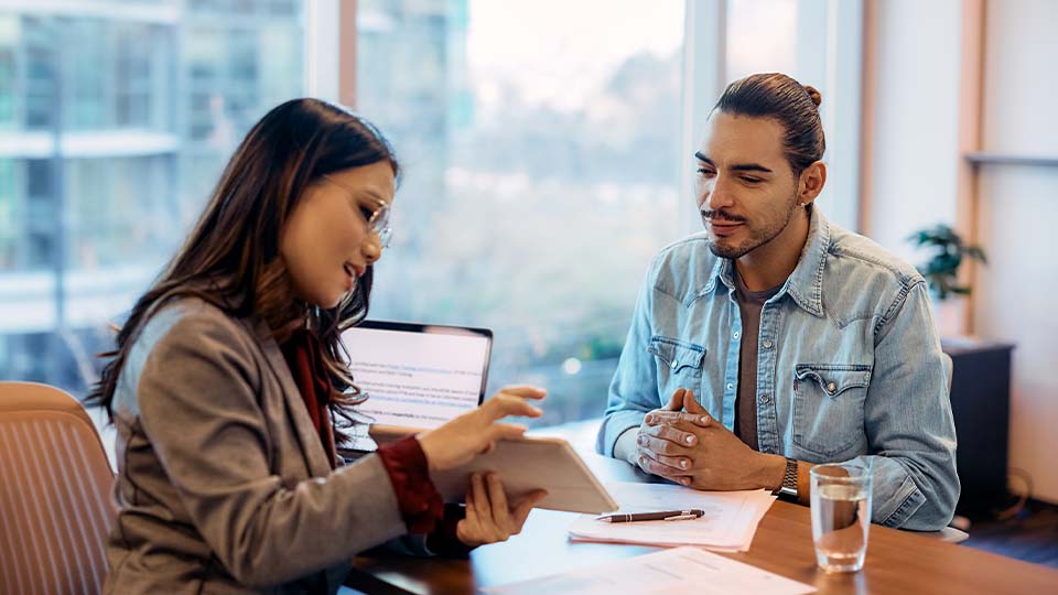 wo people are seated at a wooden table in a modern office setting with large windows in the background.