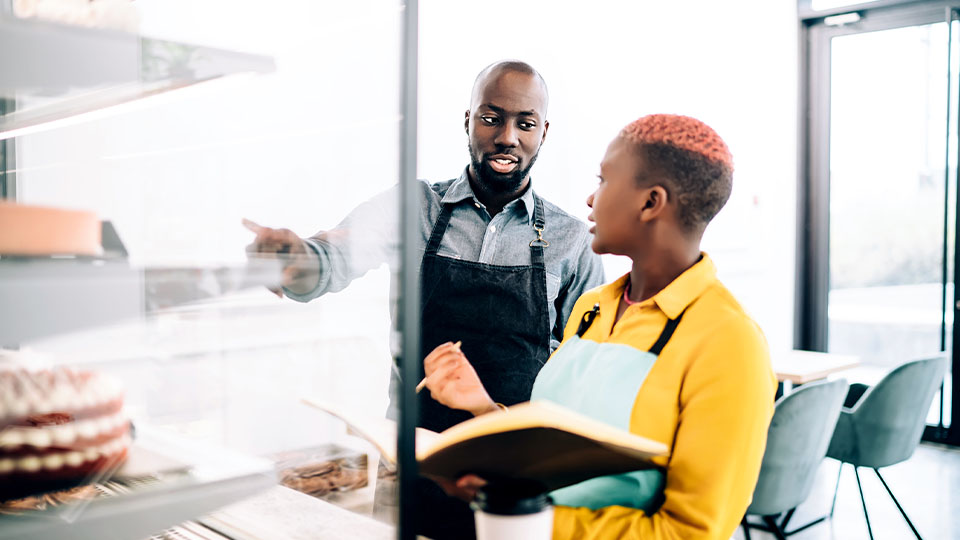 Two people wearing aprons are standing near a glass display case in what appears to be a bakery or café. One person is pointing toward items inside the display, while the other holds an open notebook.