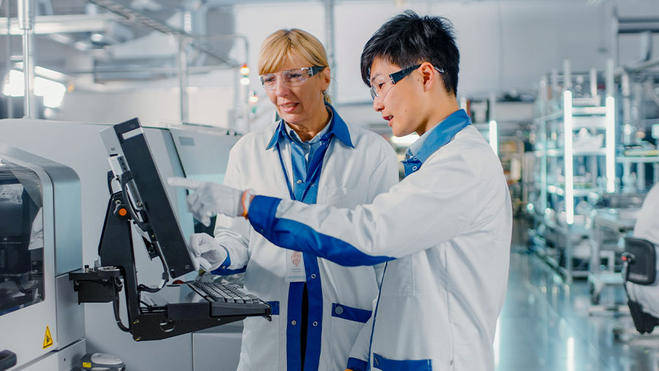 Two individuals in lab coats and protective eyewear work on a computer in a semiconductor production facility.