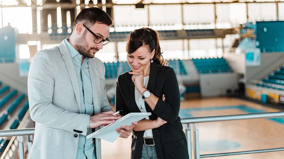 Two people are standing in an indoor sports arena near a railing, reviewing information on a tablet. The background shows rows of blue seats and a basketball court with visible markings.