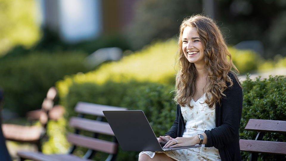 Wide angle of girl with laptop sitting on outdoor bench smiling in the sunshine