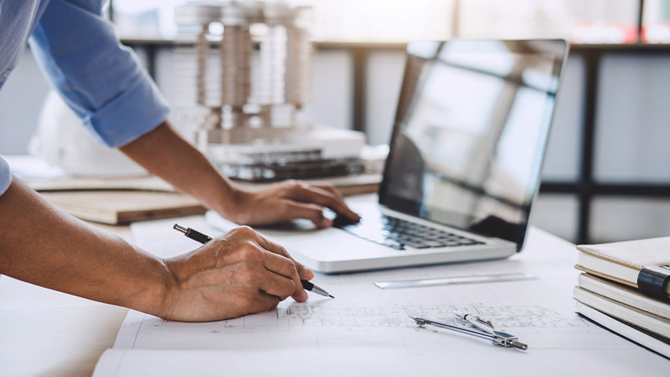 Close-up of a person's hands working on engineering plans with a laptop and design tools on the desk
