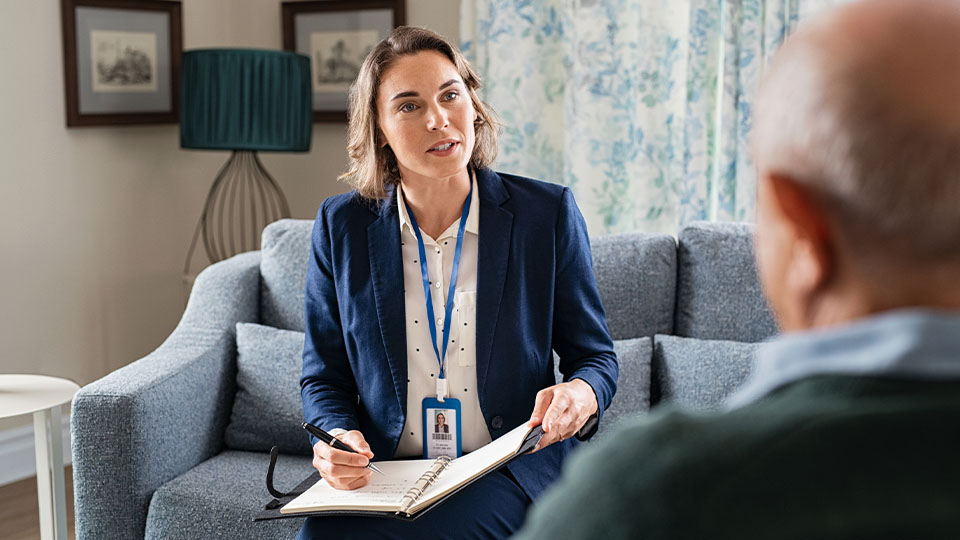 A medical professional sitting on a couch holds a pen and notebook while in conversation with a client.