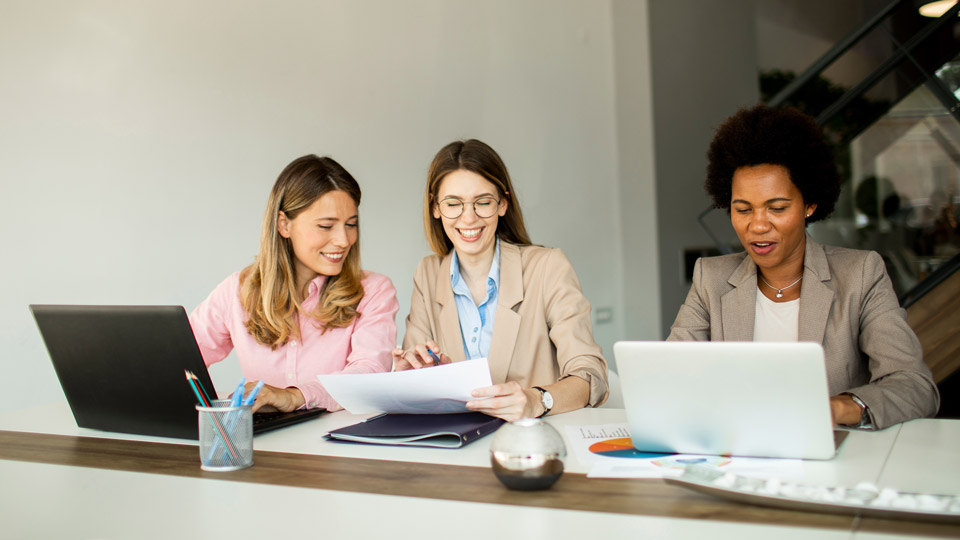 Three women working together at a desk with laptops and documents spread out. Two women are looking at papers, and the third woman is using a laptop