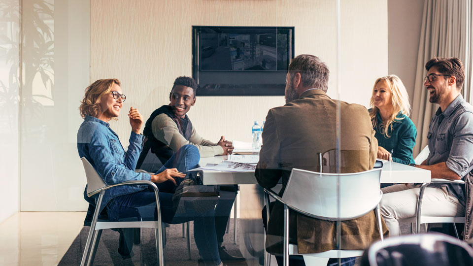 A group of five people having a meeting around a rectangular table in a modern, glass-walled conference room